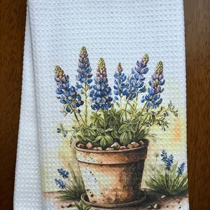 May include: White kitchen towel with a waffle weave texture. The towel features a watercolor illustration of bluebonnet flowers in a brown pot. The flowers have blue and purple petals and green leaves.