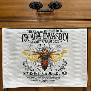 May include: White kitchen towel with a detailed illustration of a cicada and the text "CICADA INVASION SUMMER SCREAM 2024." The towel also lists states. The cicada has yellow and brown wings.