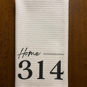 以下が含まれることがあります： テクスチャ加工された表面の白いキッチンタオル。「Home」の文字が筆記体で、その下に数字の「314」が太字で書かれています。タオルは長方形で折りたたまれています。