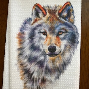 Puede incluir: Toalla blanca con una ilustración detallada de la cara de un lobo. El lobo tiene una mezcla de pelaje gris, marrón y azul, con ojos y orejas naranjas. La toalla está doblada y tiene una superficie texturizada.