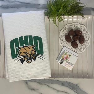 May include: White waffle-weave kitchen towel featuring the word "OHIO" in green and a green and gold bobcat logo. The towel is on a white tray with chocolates and a small card that says "Thinking of You".