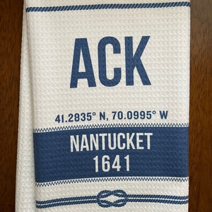 May include: White towel with navy blue accents and text. The word "ACK" is prominently displayed in large navy blue letters. Below, it reads "41.2835° N, 70.0995° W", "NANTUCKET", and "1641". A nautical knot design is at the bottom.