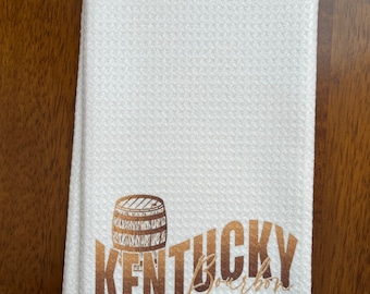 May include: White kitchen towel with a textured pattern. The towel features a brown illustration of a barrel and the words "Kentucky Bourbon" in a gradient brown font.