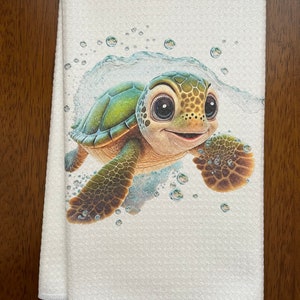 May include: White towel featuring a smiling cartoon sea turtle with a green shell and brown flippers. The turtle is surrounded by bubbles and water, creating a playful, aquatic theme. The towel is folded and ready for use.