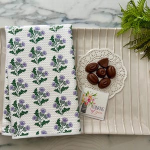 Könnte beinhalten: Ein weißes rechteckiges Serviertablett mit Streifenmuster enthält ein gefaltetes weißes Handtuch mit einem lila Distelmuster, Pralinen und eine Karte mit der Aufschrift "Thinking of you". Ein kleiner Farn befindet sich in der oberen rechten Ecke.