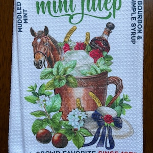 May include: White cotton kitchen towel with a colorful illustration of a mint julep cocktail. The design includes a horse, bourbon bottle, mint sprigs, and the text "Mint Julep". The towel also features the text "A Crowd Favorite Since 1875".