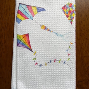 Puede incluir: Toalla de cocina blanca con textura de gofre, con ilustraciones de acuarela coloridas de cometas. Las cometas son de varias formas y colores, con tonos arcoíris, azules, amarillos y rojos. Una guirnalda de pequeñas banderas decora la parte inferior.