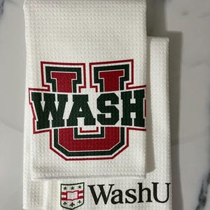 May include: White waffle-weave towels featuring the text "WASH" in a red and green block letter design. A smaller towel below has the text "WashU" with a shield and star graphic. The towels are folded and stacked.