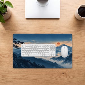 Könnte beinhalten: Eine rechteckige Schreibtischunterlage mit einer gedruckten Landschaftsillustration von Gebirgen, Kiefern und einer untergehenden Sonne in Blau-, Orange- und Weißtönen. Eine weiße Computertastatur und eine weiße Computermaus liegen auf der Unterlage.