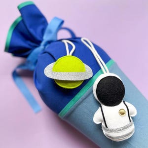Pu&ograve; includere: Un sacco blu e verde con coulisse e ornamenti di feltro a forma di pianeta e astronauta. Il pianeta &egrave; verde lime con un anello di glitter argentati. L'astronauta &egrave; bianco con un casco nero e dettagli argentati.