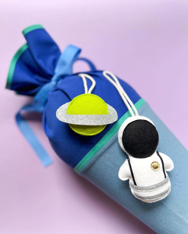 Pu&ograve; includere: Un sacco blu e verde con coulisse e ornamenti di feltro a forma di pianeta e astronauta. Il pianeta &egrave; verde lime con un anello di glitter argentati. L'astronauta &egrave; bianco con un casco nero e dettagli argentati.