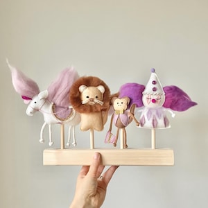May include: A whimsical display of handmade felt animal figures, including a white horse with pink wings, a lion, a monkey, and a clown. The figures are mounted on wooden dowels and a wooden base, showcasing a soft color palette.