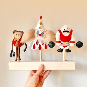 Könnte beinhalten: Ein Set aus drei Filz-Zirkusfiguren auf einem Holzständer. Die Figuren umfassen einen Clown mit einem weißen und roten Kleid, einen Affen mit einer roten Jacke und einer Fliege und einen starken Mann mit einem roten und schwarzen Kostüm, der eine Hantel hebt.