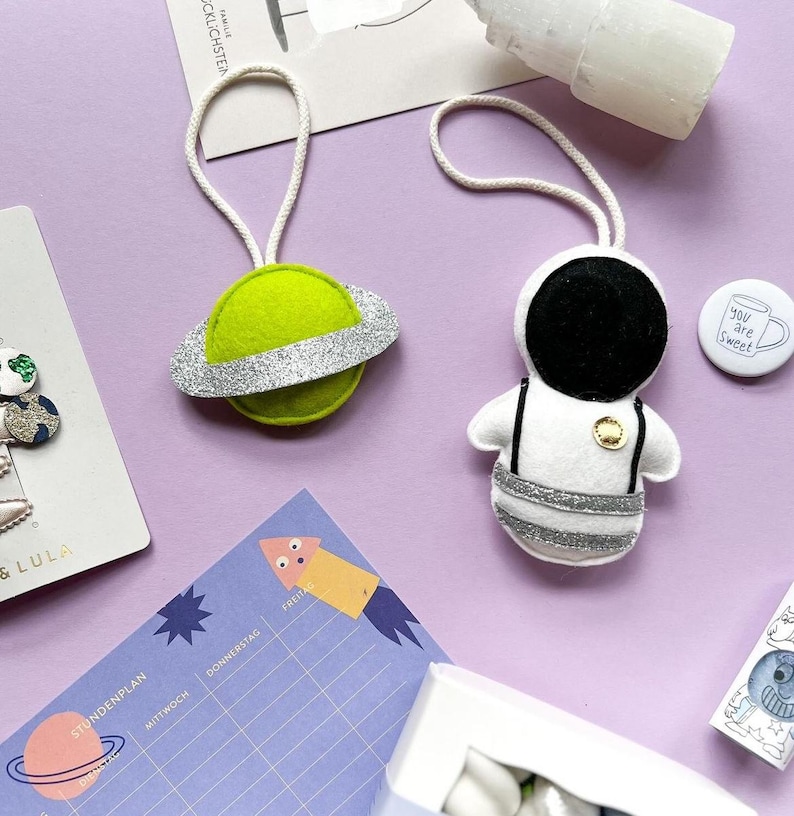 Pu&ograve; includere: Ornamenti in feltro a tema spazio. Un pianeta verde con un anello glitterato argentato e un astronauta bianco e nero. &Egrave; visibile anche un bottone con la scritta "You are Sweet", oltre ad altri articoli di cancelleria.