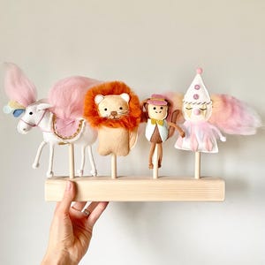 May include: A set of five felt animal puppets on wooden sticks. The puppets include a unicorn, a lion, a monkey, and a clown. The puppets are arranged on a wooden base.