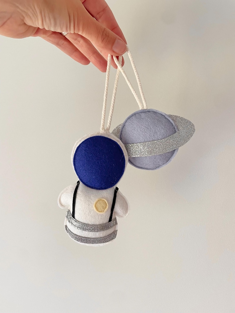 Pu&ograve; includere: Due ornamenti in feltro a tema spaziale. Uno &egrave; un astronauta con casco blu e dettagli argentati. L'altro &egrave; un pianeta viola chiaro con un anello di glitter argentati. Entrambi hanno cordini bianchi.