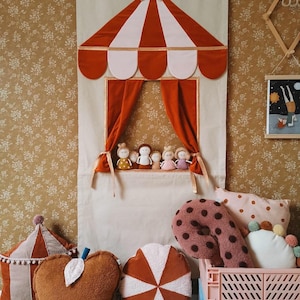 May include: A red and white circus tent puppet stage with a gold star on top. The stage has a red velvet curtain and is decorated with small dolls. There are several plush pillows in the foreground, including a brown apple-shaped pillow, a brown and white striped pillow, and a brown and pink polka dot pillow.