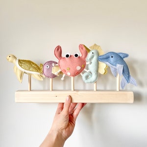Op de afbeelding: Een houten plank met vijf vilt zeedieren op stokjes. De dieren zijn een schildpad, een krab, een zeepaardje, een vis en een dolfijn. De plank is geschilderd in een lichte houtkleur.