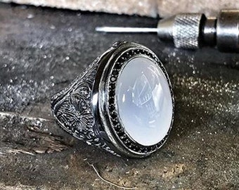 May include: A silver ring with an oval-shaped white stone set in a black bezel. The ring band is intricately engraved with a floral design.