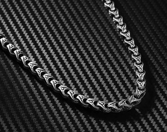Könnte beinhalten: Eine silberne Kettenkette mit einem detaillierten, verzierten Muster. Die Kette liegt auf einem schwarzen, strukturierten Hintergrund.
