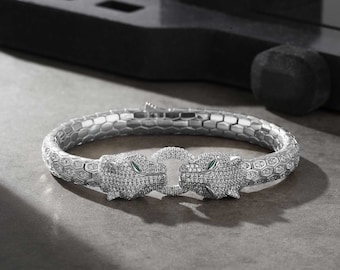 May include: A silver bracelet with a textured design and two panther heads on the clasp. The panther heads are encrusted with clear and green gemstones.