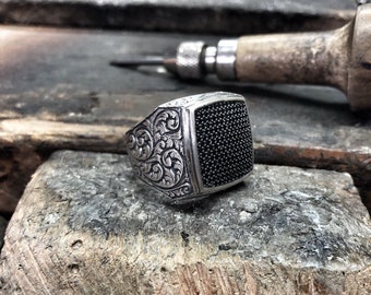 May include: A silver ring with a black square stone set in the center. The ring is decorated with an intricate floral design.