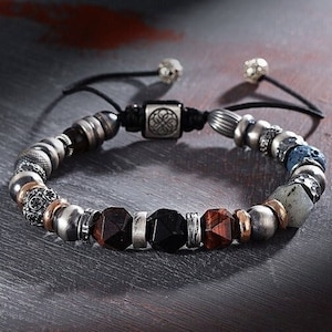 May include: A beaded bracelet with a variety of materials, including wood, metal, and stone. The bracelet features a silver charm with a knot design and a black cord.
