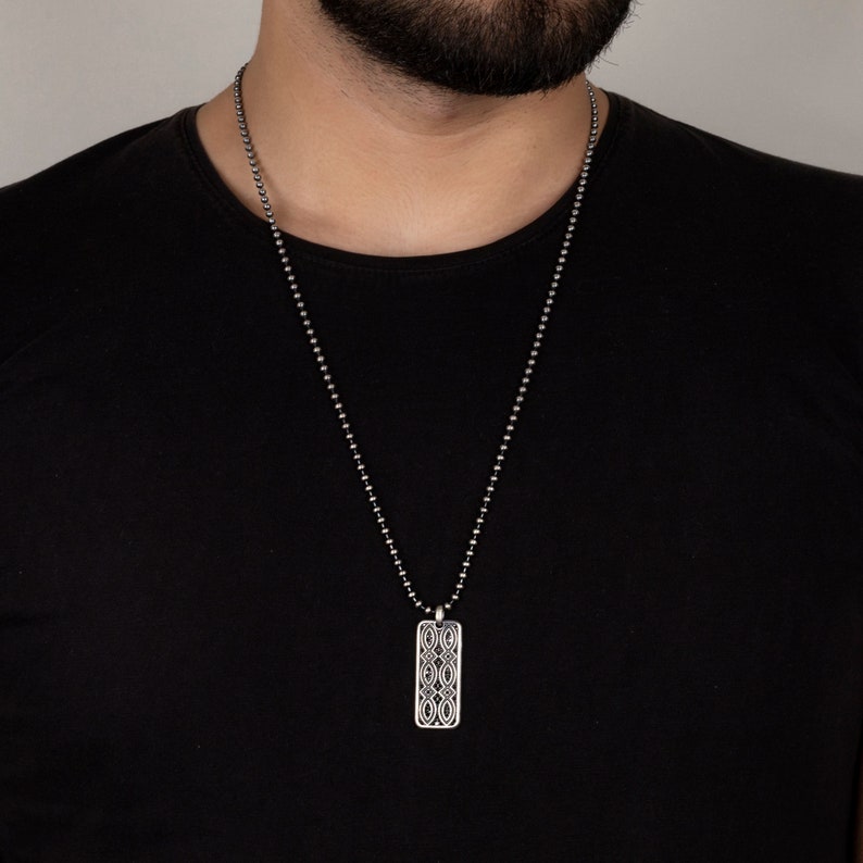 Puede incluir: Un collar de cadena de plata con un colgante de plata con un dise&ntilde;o geom&eacute;trico. El colgante es de forma rectangular y tiene un patr&oacute;n detallado.