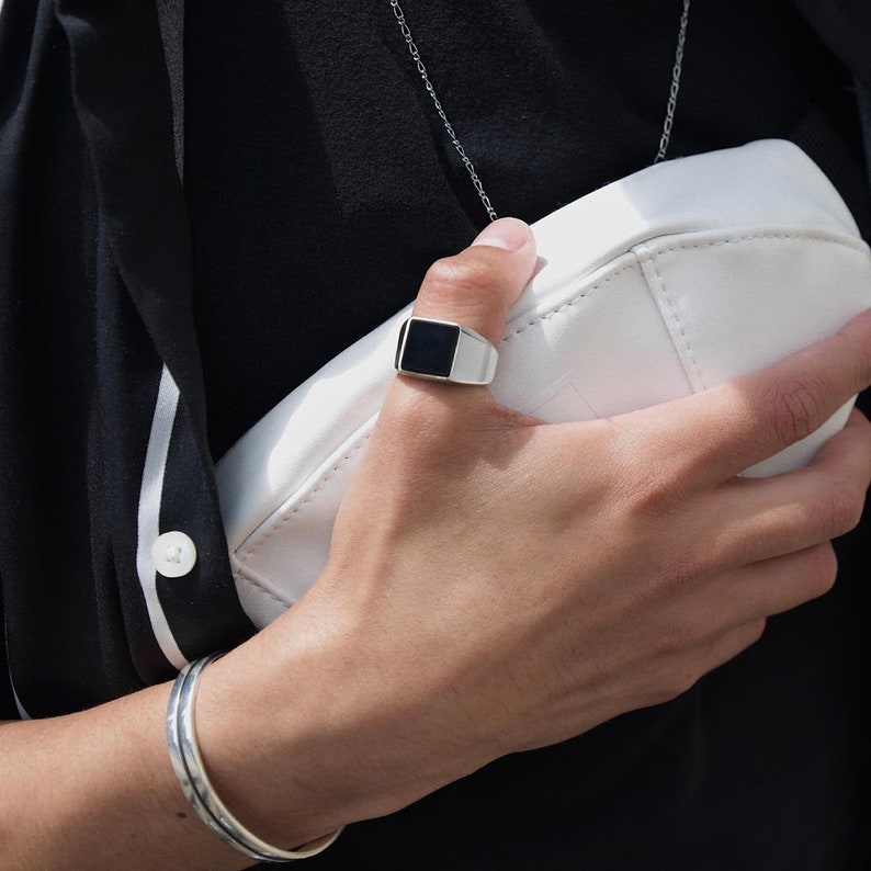 May include: A silver ring with a black square stone on the top. The ring is worn on the thumb of a person's hand.