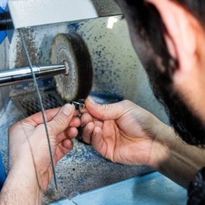 May include: A person is polishing a metal object with a buffing wheel. The object is held in their left hand and the buffing wheel is spinning in a clear plastic enclosure.