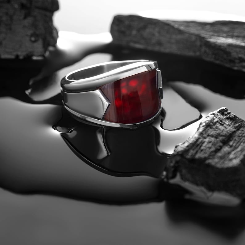 May include: A silver ring with a large, red gemstone set in the center. The ring is sitting on a black surface with pieces of charcoal around it.