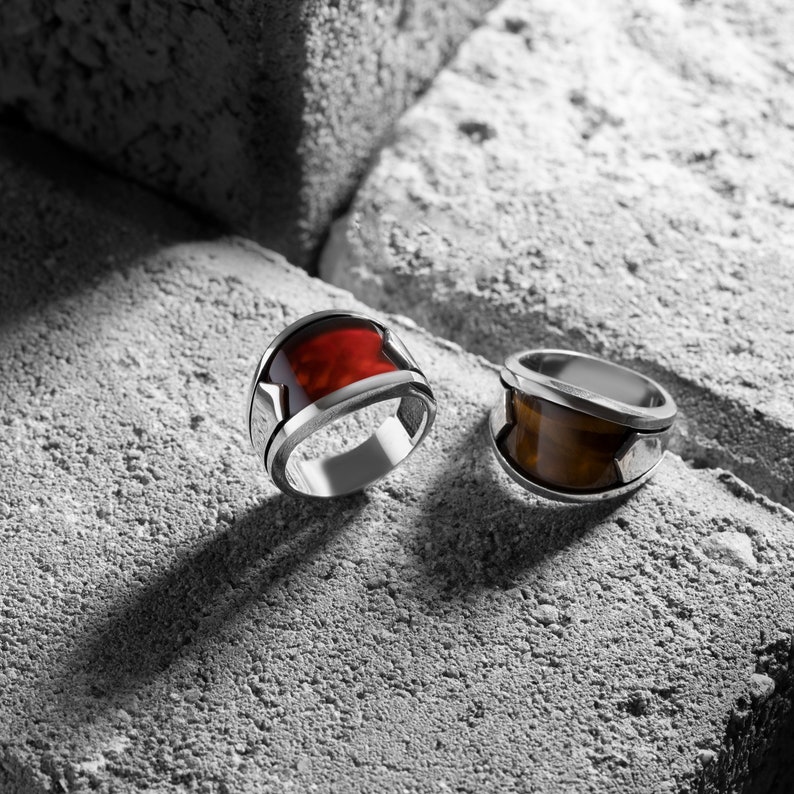 May include: Two silver rings with red and brown gemstones set in the center. The rings are on a gray textured surface.