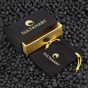 Puede incluir: Una caja negra con ribetes dorados y el texto "SULTANART" en letras doradas. La caja est&aacute; abierta y una bolsa de terciopelo negro con el mismo logotipo est&aacute; dentro.