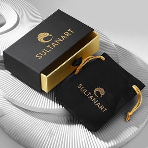May include: A black box with gold trim and the text "SULTANART" on the top. The box is open and a black velvet pouch with the same text is inside.