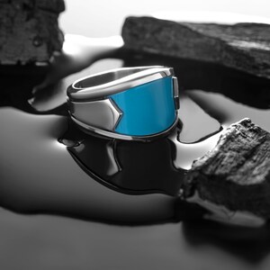 May include: A silver ring with a blue stone set in the center. The ring is sitting on a black surface.