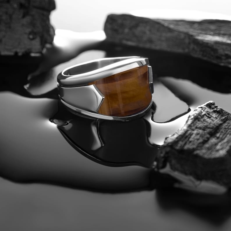 May include: A silver ring with a brown tiger's eye stone set in the center. The ring is sitting on a black surface with a reflection of the ring in the surface.