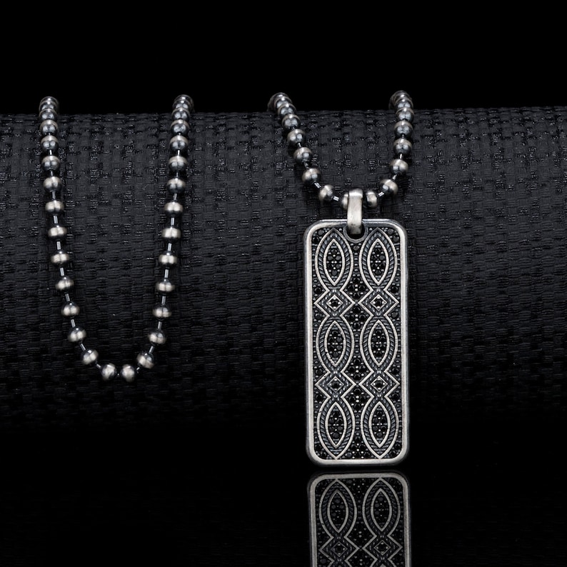 Puede incluir: Un collar de cadena de plata con un colgante rectangular con un dise&ntilde;o geom&eacute;trico negro.