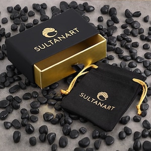 May include: A black box with a gold interior and the text "SULTANART" on the top. The box is open and a black velvet pouch with the same logo is visible. The pouch is on a background of black pebbles.