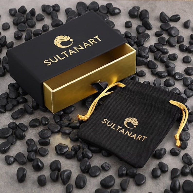 May include: A black box with a gold interior and the text "SULTANART" on the top. The box is open and a black velvet pouch with the same logo is visible. The pouch is on a background of black pebbles.