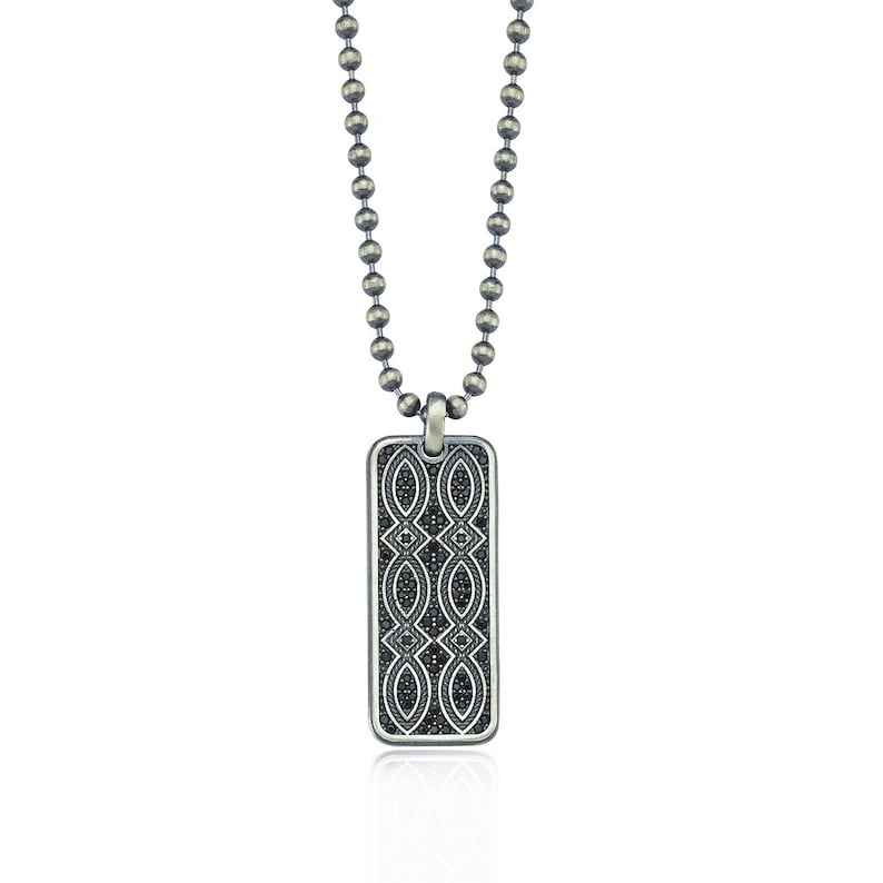 Puede incluir: Un collar de cadena de plata con un colgante rectangular de plata. El colgante est&aacute; decorado con un patr&oacute;n geom&eacute;trico negro.