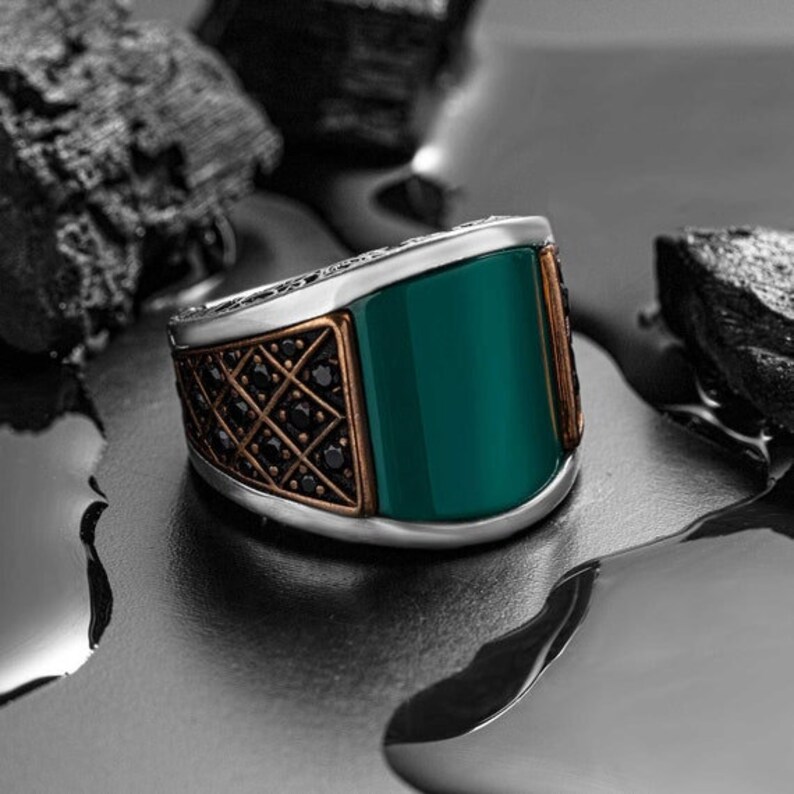 May include: A silver ring with a green stone and black accents. The ring has a geometric design and is set in a silver band.