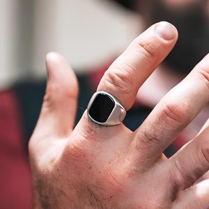 Könnte beinhalten: Ein silberner Ring mit einem schwarzen rechteckigen Stein.