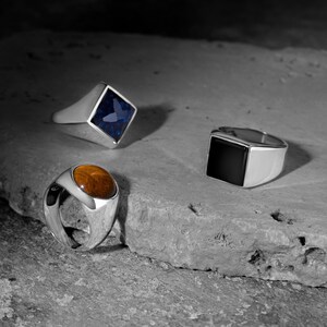 May include: Three silver signet rings with different coloured gemstones. The first ring has a blue gemstone, the second ring has a brown gemstone, and the third ring has a black gemstone.