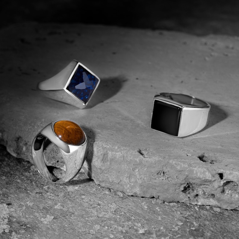May include: Three silver signet rings with different coloured gemstones. The first ring has a blue gemstone, the second ring has a brown gemstone, and the third ring has a black gemstone.