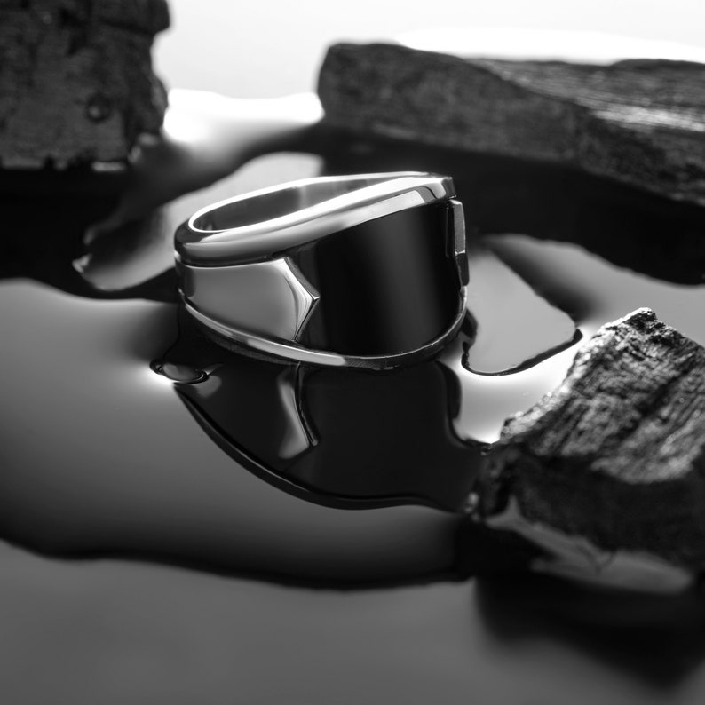 May include: A silver ring with a black stone set in a geometric design. The ring is lying on a black surface with pieces of charcoal.