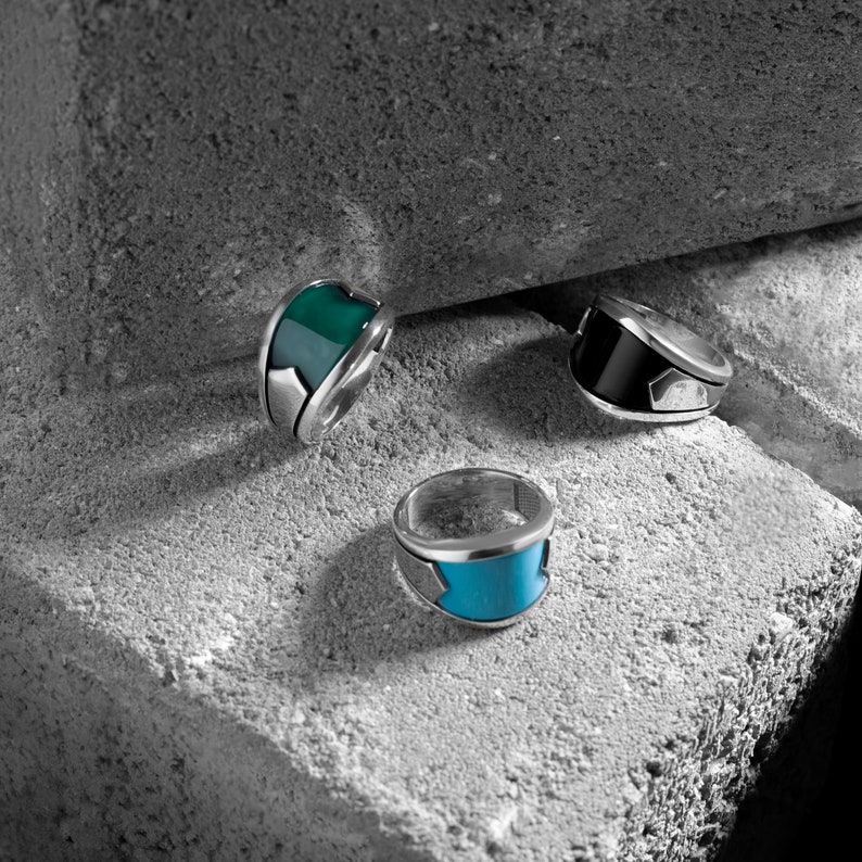 May include: Three silver rings with colorful inlays. The rings have a silver band with a green, blue, and black inlay. The rings are on a gray textured surface.