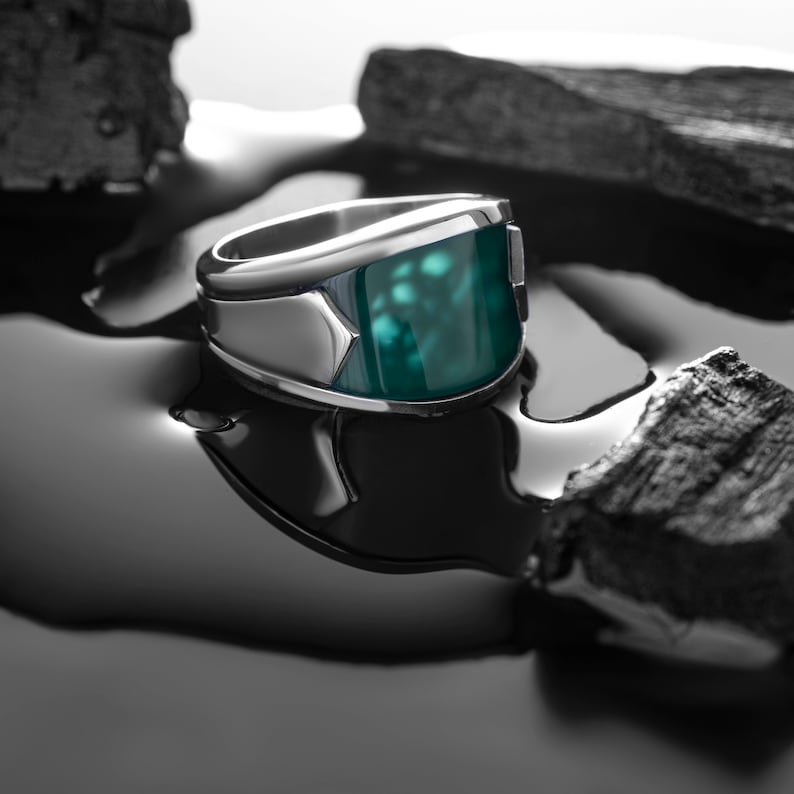 May include: A silver ring with a large, green gemstone set in a square bezel. The ring is sitting on a black surface.