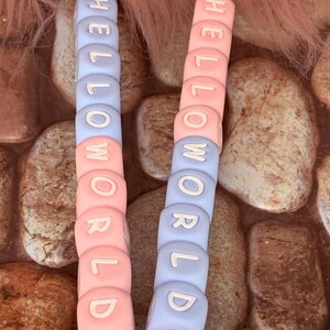 May include: Two baby pacifier clips with the words "HELLO WORLD" spelled out in white letters on pink and blue beads. Each clip has a silver clasp and a pink ribbon.