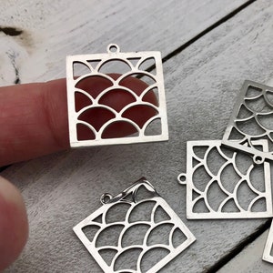 May include: Several silver-colored square charms with a cut-out scallop design. Each charm has a small loop at the top for attaching to jewelry. The charms are scattered on a light-colored wooden surface.