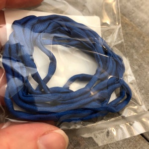 May include: A bundle of blue fabric cord, tightly coiled within a clear plastic bag. The cord transitions from a lighter shade of blue to a deeper, richer hue. The texture of the fabric appears soft and pliable.